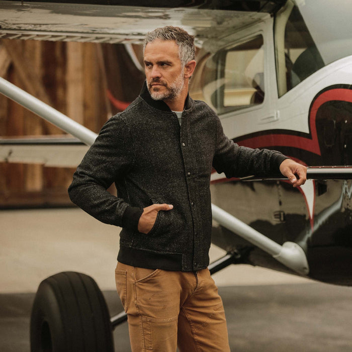 fit model wearing The Bomber Jacket in Charcoal Wool leaning against an airplane